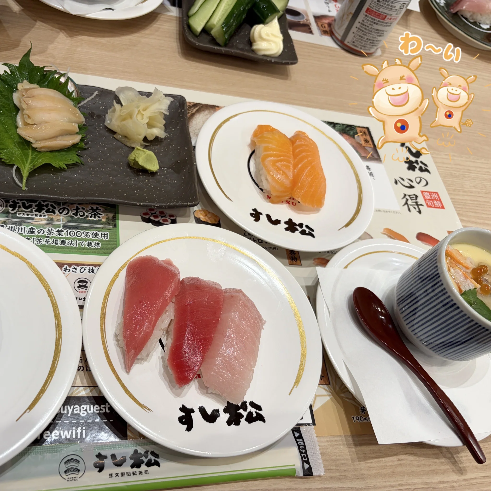 すし松 三鷹南口店の食事ショット。手前にマグロ赤身2貫の白いすし松ロゴ皿、奥にサーモン2貫の白いすし松ロゴ皿、左にがり・わさび・大葉、右にきゅうり、右下に黒い茶碗の茶碗蒸し、左奥に『すし松のお茶 三川川産の茶葉100%使用』のリーフレット、右上にマッキー&ヤッキーの『わ〜い』オーバーレイ