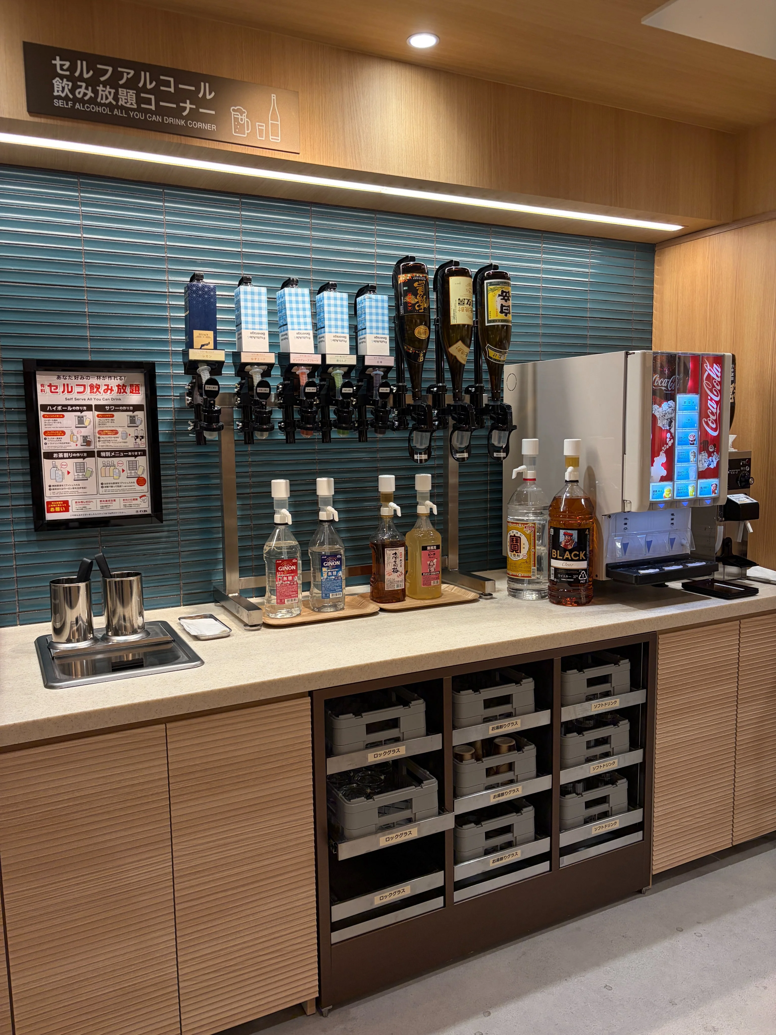 すし松 三鷹南口店のセルフアルコール飲み放題コーナー。日本語表記『セルフアルコール飲み放題コーナー』英語表記『SELF ALCOHOL ALL-YOU-CAN-DRINK CORNER』。左に4基のビールサーバー（青ハンドル）、隣に4本のハイボール・ウイスキーサーバー、カウンター上に焼酎・梅酒・リキュールのボトル、右にソフトドリンクサーバー、下段はビール瓶ケース