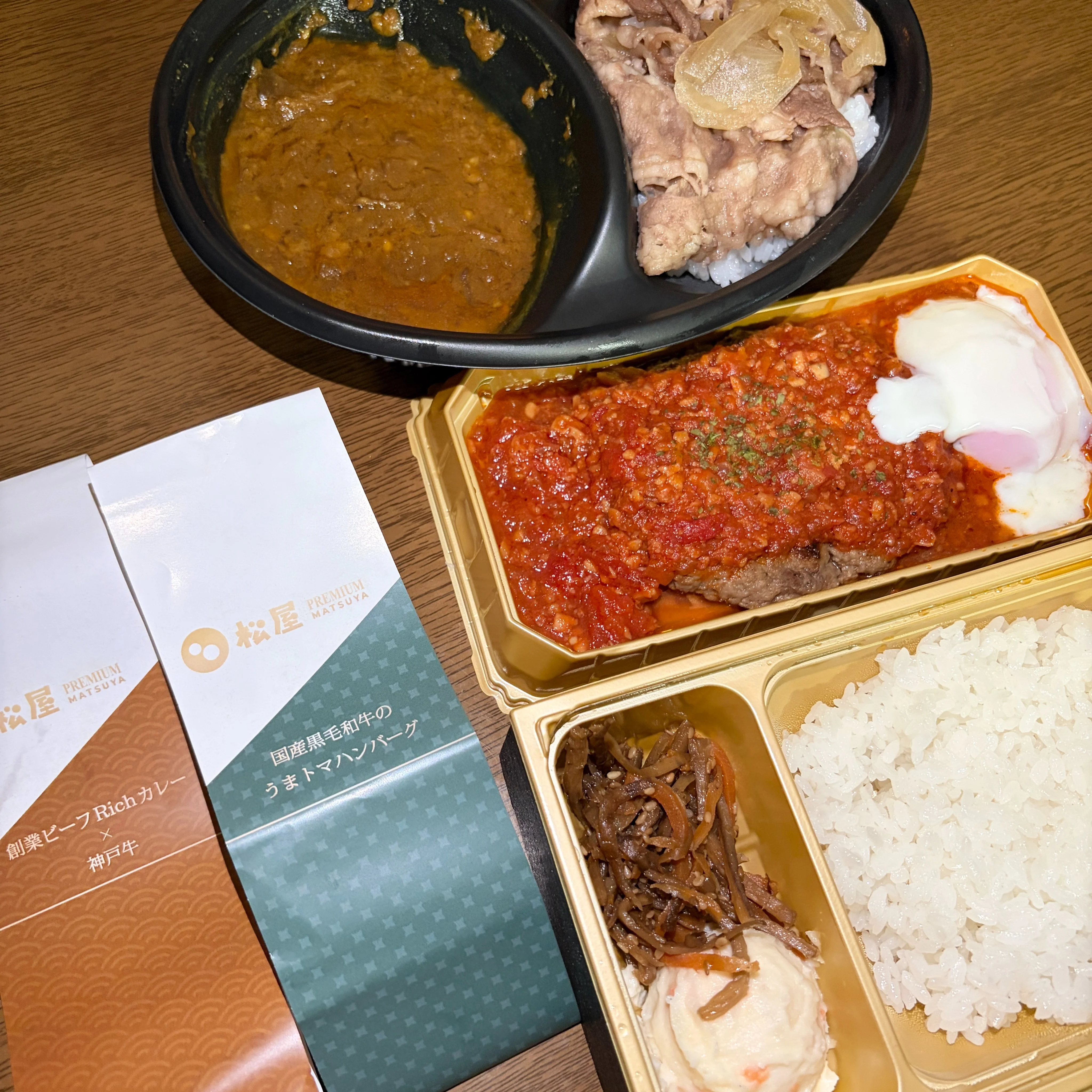 松屋PREMIUM 銀座店2品の家族喫食。上部に創業ビーフRichカレー×神戸牛の黒い丸容器、下部に国産黒毛和牛のうまトマハンバーグの金色四角弁当容器、左に商品パッケージスリーブ2点が立てかけられている