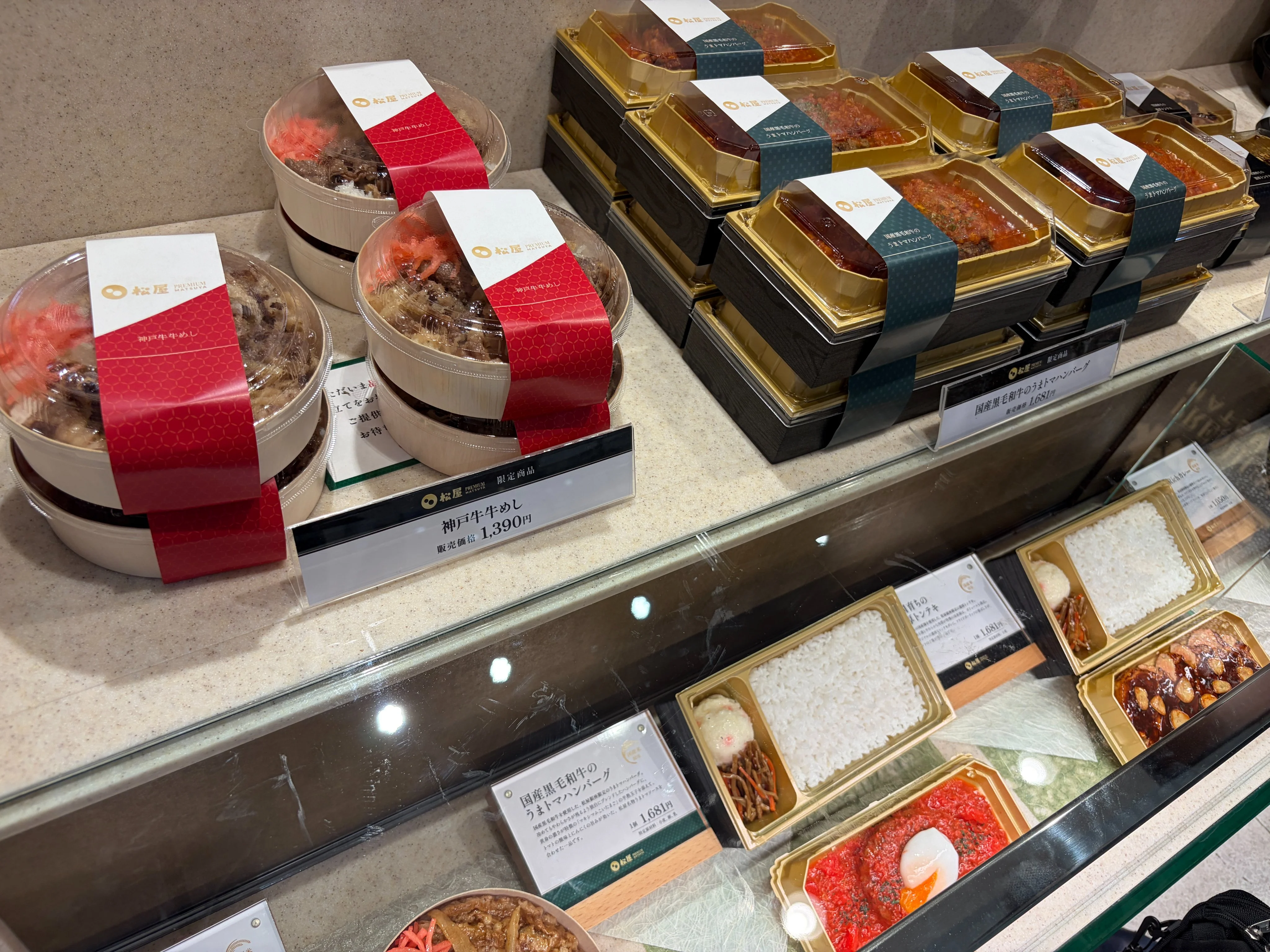 松屋PREMIUM 銀座店ガラスケース内の商品陳列。上段左に赤帯付きの『神戸牛 牛めし 1,390円』丸容器、上段右に金色のテイクアウト容器が並ぶ